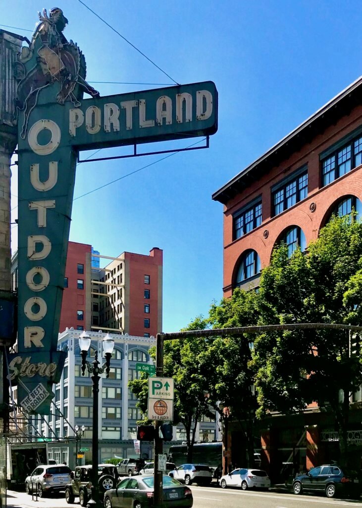 The iconic Portland Outdoor Store in downtown Portland shows off a green retro style neon sign with white lettering while other vintage red brick buildings stand across the street and further down.