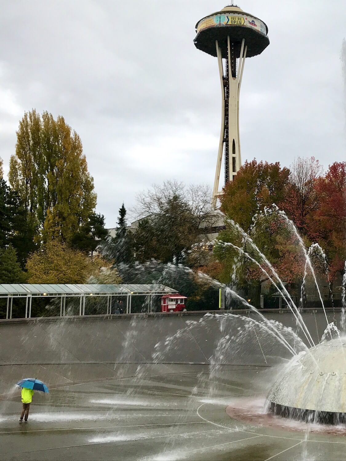 A boy wearing a yellow rain jacket approaches the large fountain with a blue umbrella.  The metal piece of art shoots out powerful streams of water in all kinds of directions.  In the background the Seattle Space Needle rises up above the park at Seattle Center. 