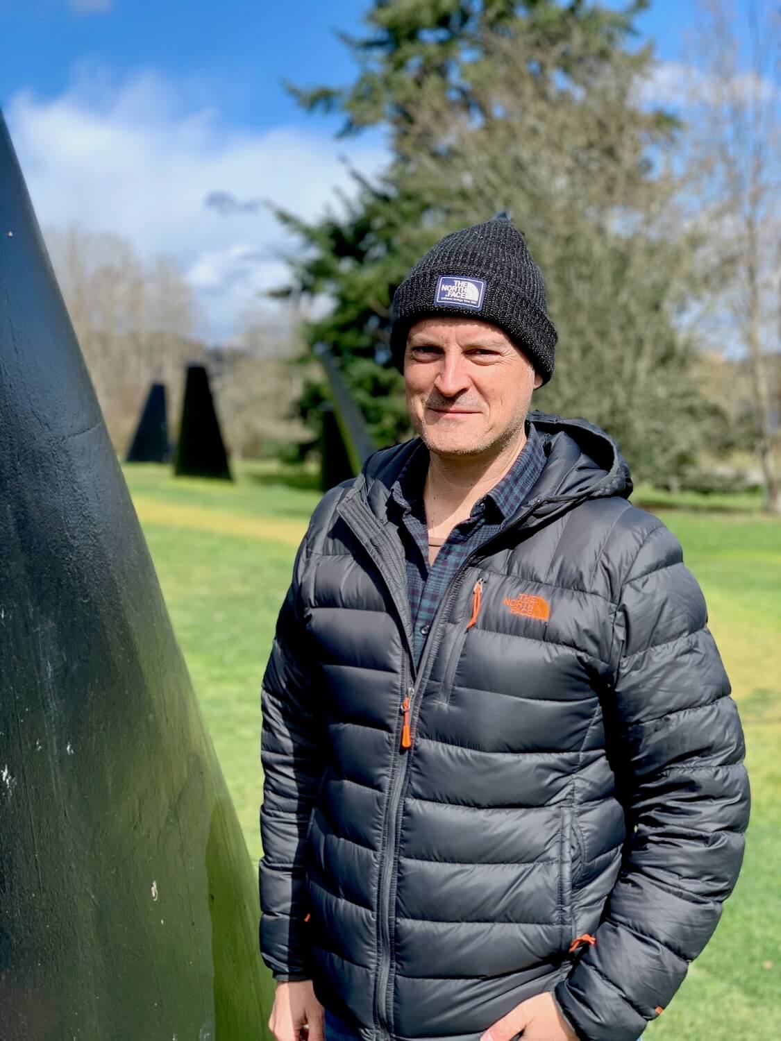 Matthew Kessi stands next to a former submarine stabilizer fin in The Fin Project.  He is wearing a North Face cap and jacket and other layers of clothing.  Other fins rise up in the out of focus background.  This is a great outdoor activity in winter in Seattle.  