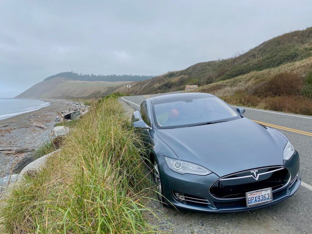 a gray Tesla sits on the side of a road just on the edge of a bank leading down to a beach on Whidbey Island in the middle of the Puget Sound
