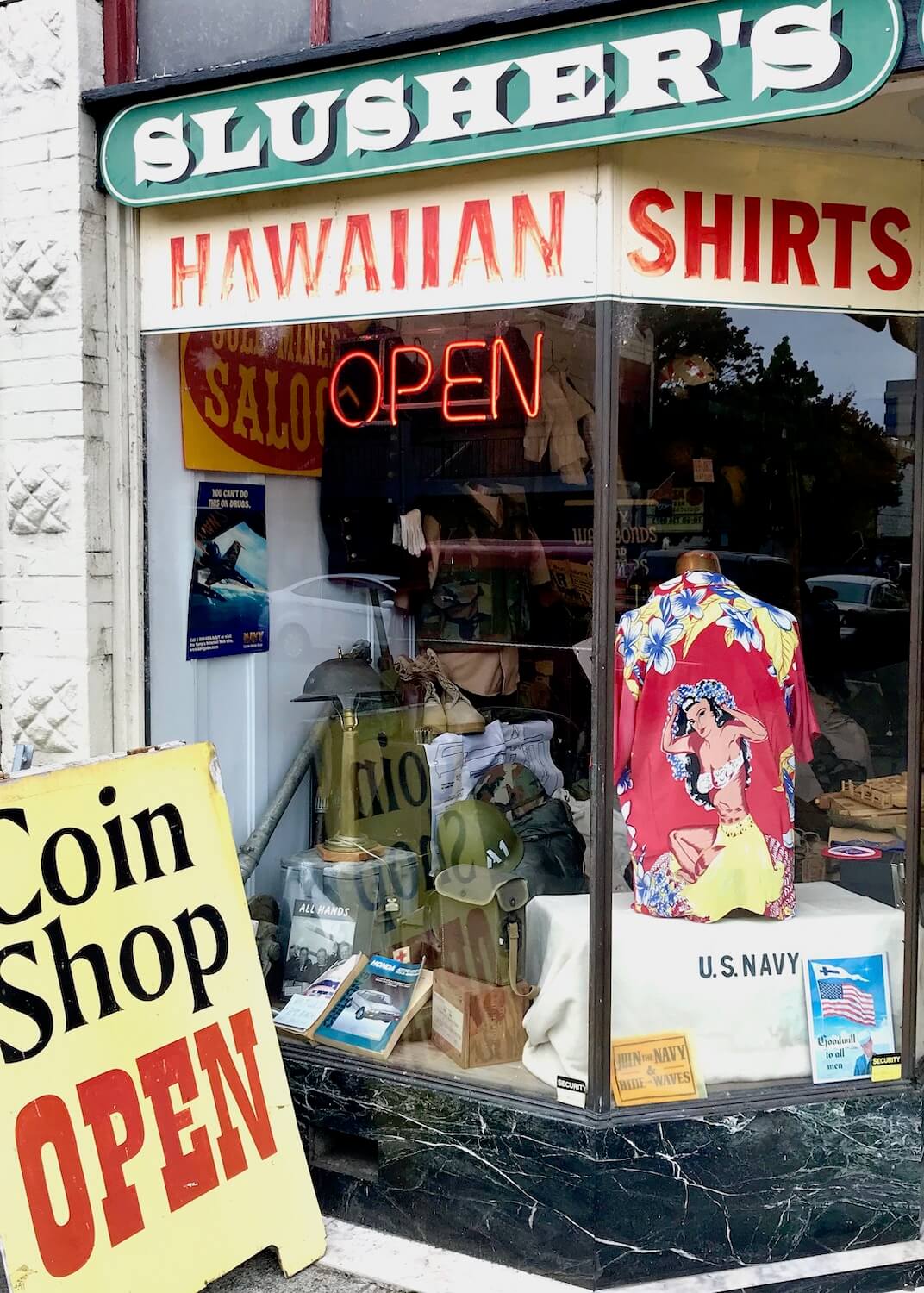 Looking through a window of an antique store in Central, Washignton which is along the Seattle to Portland drive. There is a sandwich board out front that's yellow with black and red lettering that says, "Coin Shop Open." The signage in the front window says, "Hawaiian Shirts" in red lettering. In the window there is a US Navy blanket and a Hawaiian shirt on display.