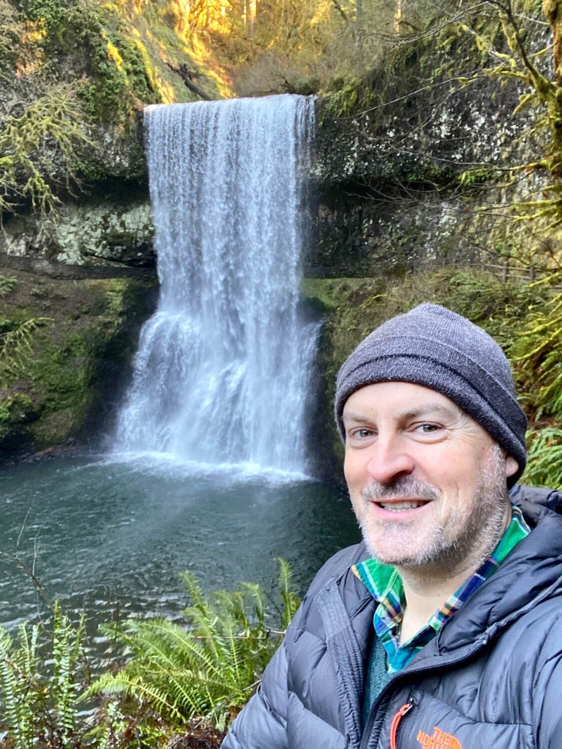 Selfie of Matthew Kessi wearing a gray wool hat and a puffy north face jacket while standing near the edge of a trail overlooking a flowing waterfall, amongst dark greenish water and surrounded dense green ferns and moss.