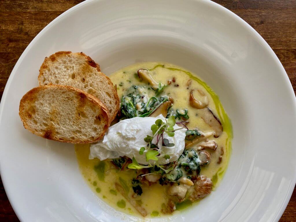 A round white porcelain bowl sits on a wooden table and contains a mixture of mushrooms in a light buttery yellow sauce with a poached egg on top. On the side of the bowl, between nine and ten o'clock are two piece of lightly toasted bruschetta.