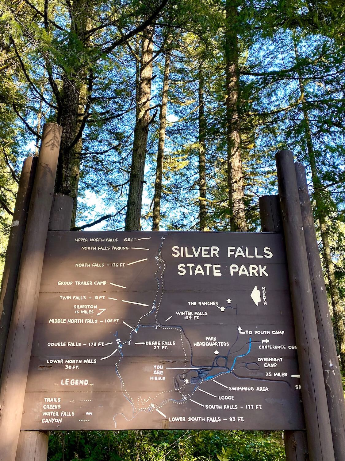 This map of Silver Falls State Park reflects the historic charm of this iconic Silver Creek Falls area.  A grouping of three logs, painted brown, support a plywood sign, also painted brown that highlights the ten waterfalls featured in the park.  The writing is in a yellow hue and outlines the various trails as well as the specific heights of the waterfalls.  In the background, mid-aged fir trees rise up to the blue sky. 