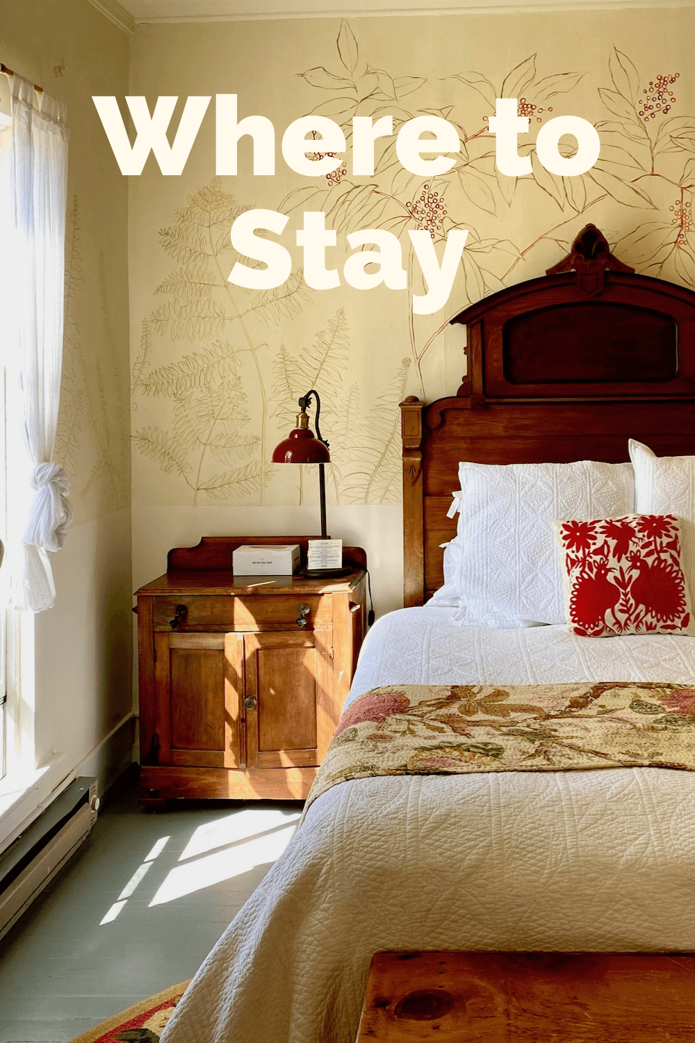 A hotel room sees sun shining through a full length window onto an antique bed frame with a cream colored wall with etched artwork.