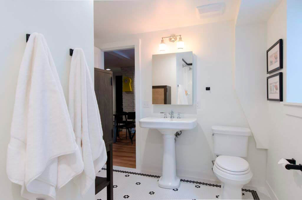 A pristine white bathroom with black accents of towel hooks and hexagon tile on the floor.  The sink is a pedestal and is next to a white toilet. 