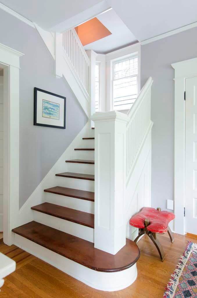 The entry to a Victorian style house in Airbnb that is used to host guests.  The stairway banister is painted white and the walls a shade of lavender in the bright summer light. 
