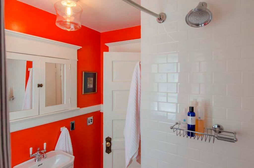 A bright reddish orange bathroom sings against white subway tile in the shower area and a brass doorknob leading out of the room.  