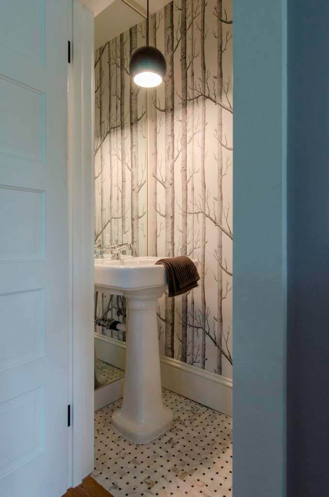 An overhead bubble like lamp shines down on a sink inside a small powder room within a home that is used as an Airbnb in Seattle, Washington.  The wallpaper is trees.  