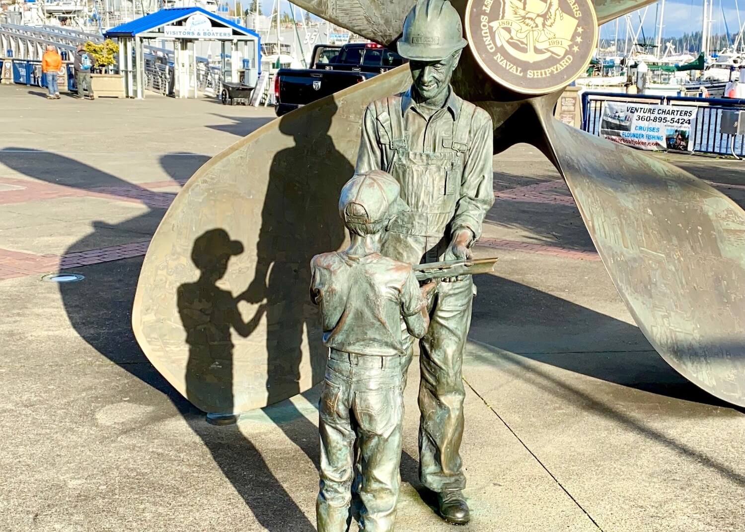 A bronze statue of a father passing on a shipbuilding tool to his young son. They are standing in front of a giant propulsion screw of a ship.