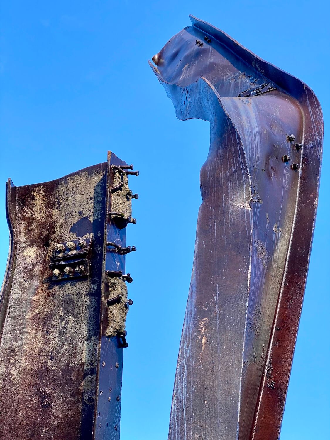 Two steel girders salvaged from Ground Zero following the attacks on the World Trade Center on 9/11.  They are purplish in color and show parts of concrete still attached to the steel and bolts loosely hanging onto the steel.  The sky in the background is a strong blue color. 