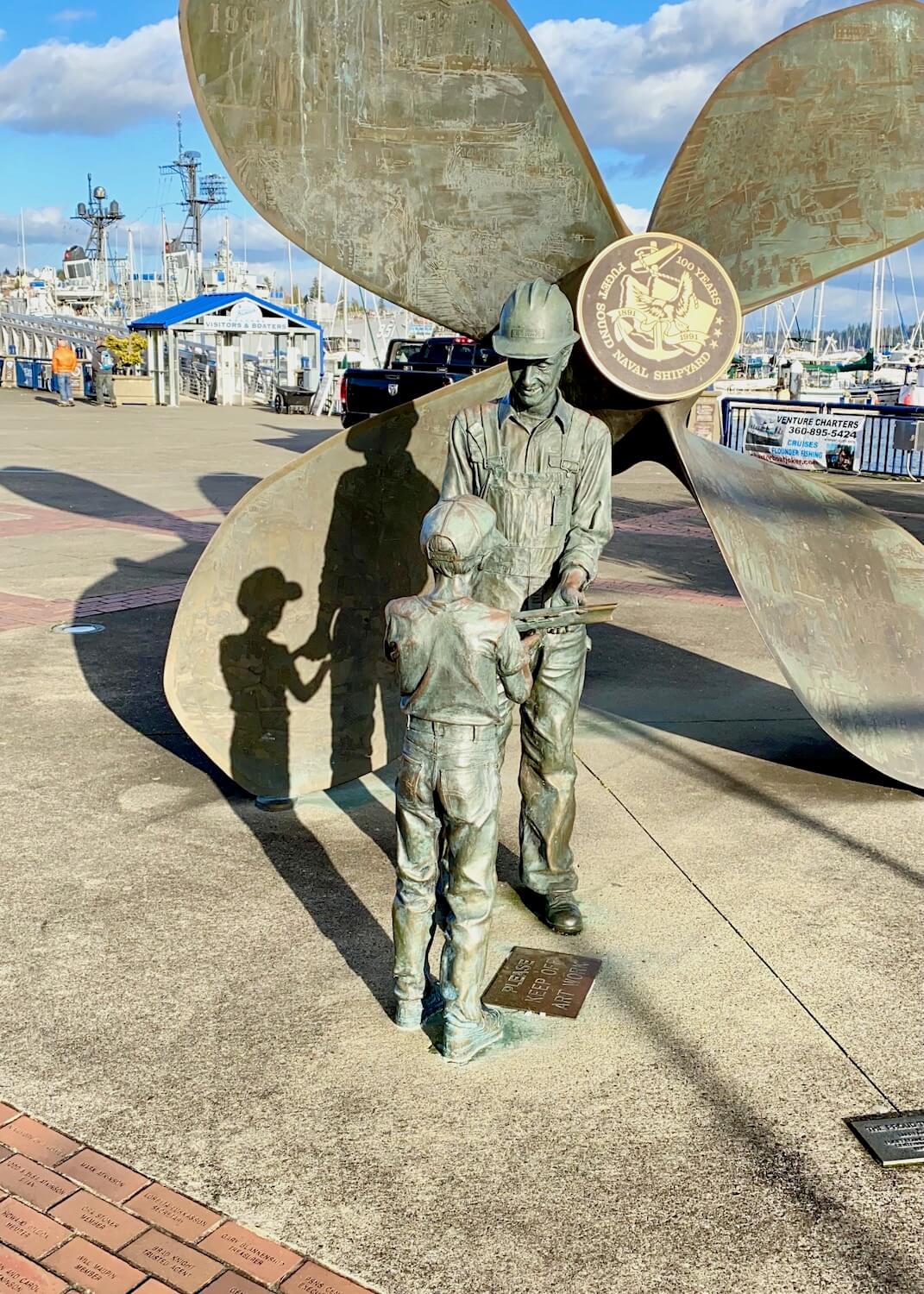 A bronze statue of a father passing on a shipbuilding tool to his young son. They are standing in front of a giant propulsion screw of a ship.