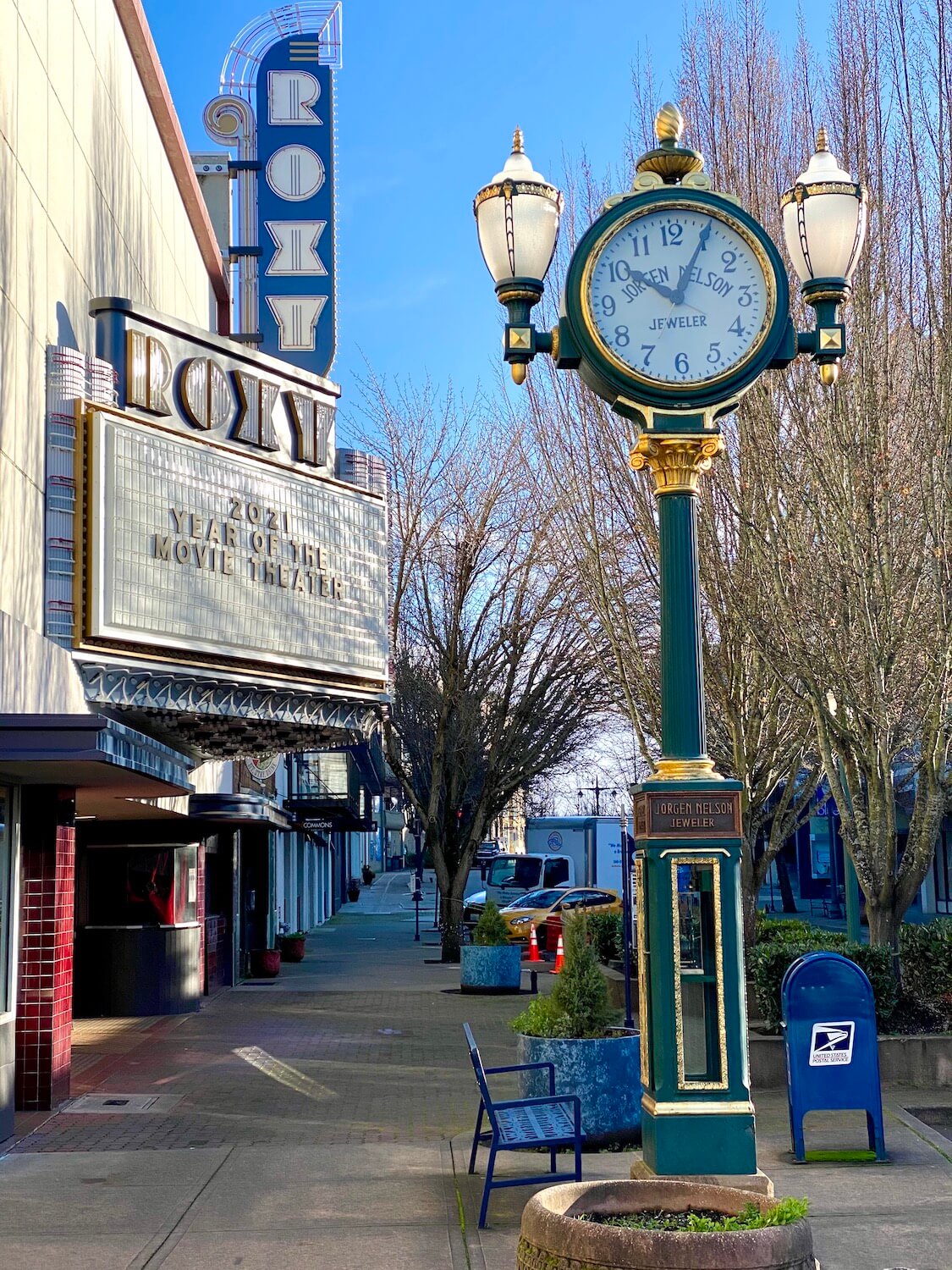 A scene in Downtown Bremerton Washington shows the Roxy movie theatre with a blue and white neon sign and an ornate sidewalk clock.  Along the sidewalk is a US Postal Service blue mailbox and the sky above is blue.