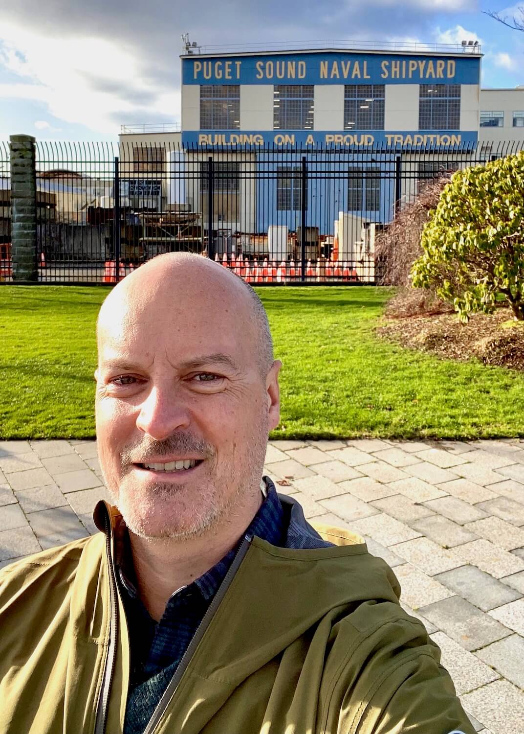 A selfie of Matthew Kessi in front of the Puget Sound Naval Shipyard in Bremerton, Washginton.  The grass is green in the background in front of a black iron grate fence leading to the industrial area. 