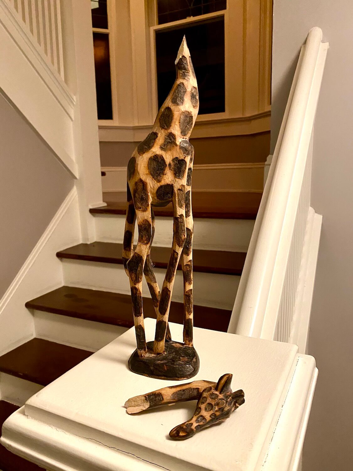 A wooden giraffe with brown spots burnt into the lighter blond wood stands on the banister of a stairway with the severed head laying down on the white banister.  