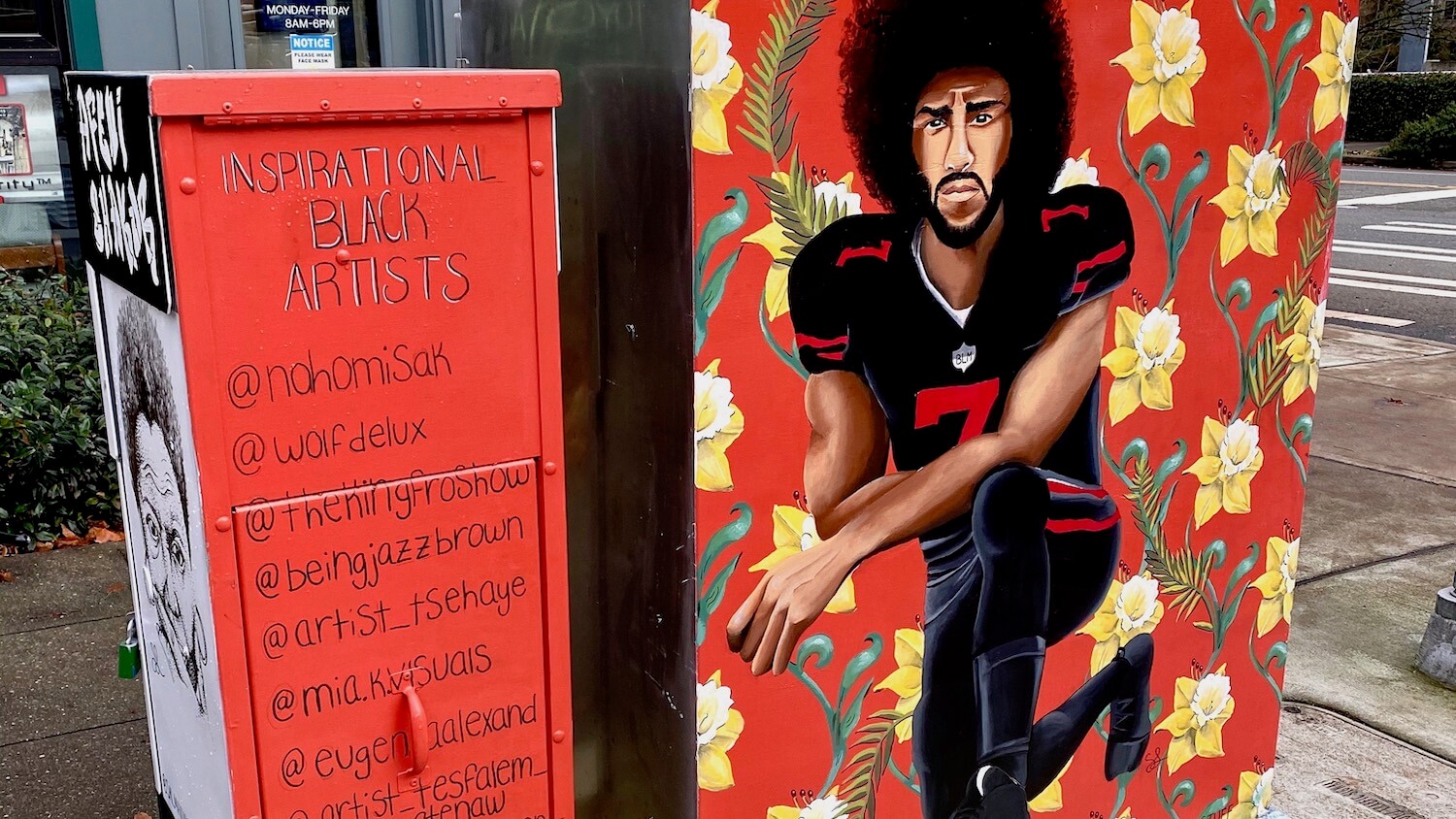 A mural of Colin Kaepernick kneeling on is pained on the side of an electrical box in Seattle.  His football uniform is black with red lettering and he is surrounded by yellow daffodils  and a red background, that continues to a different electrical box with a list of inspirational black artists.  