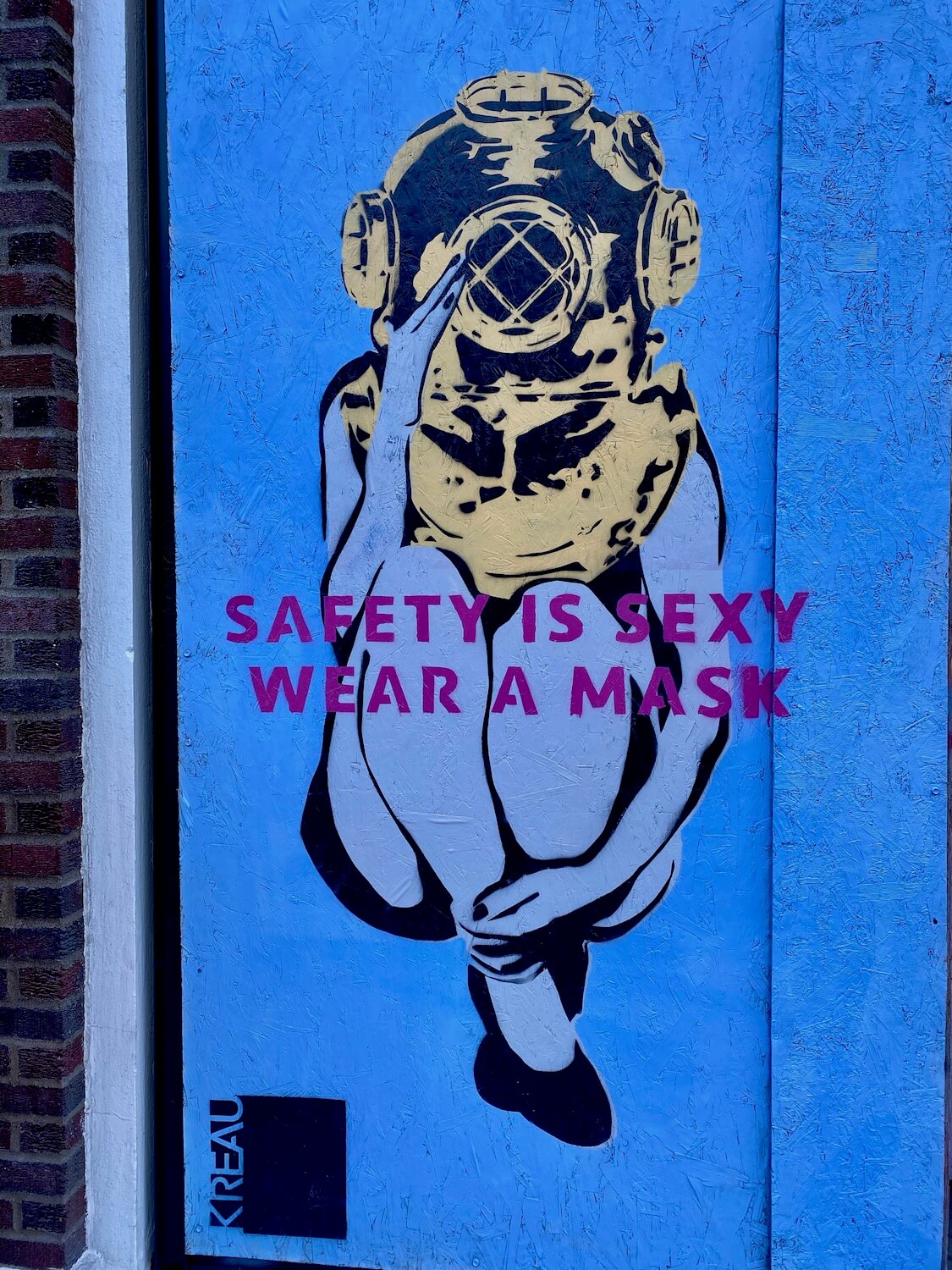 This mural painted on a piece of plywood covering a store front shows a woman sitting down wearing an old fashioned scuba diving metal helmet.  The background is blue and lettering in purple says, "Safety is sexy.  Wear a mask."  