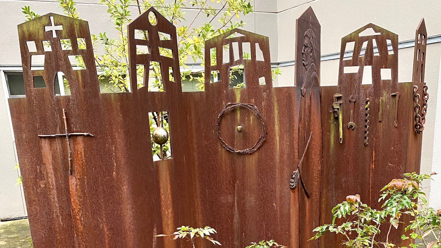 A rusted metal sculpture stands upright on the campus of Seattle University with six main spires that appear to be buildings and symbolic pieces below range from a chain to a cross made from twigs. 