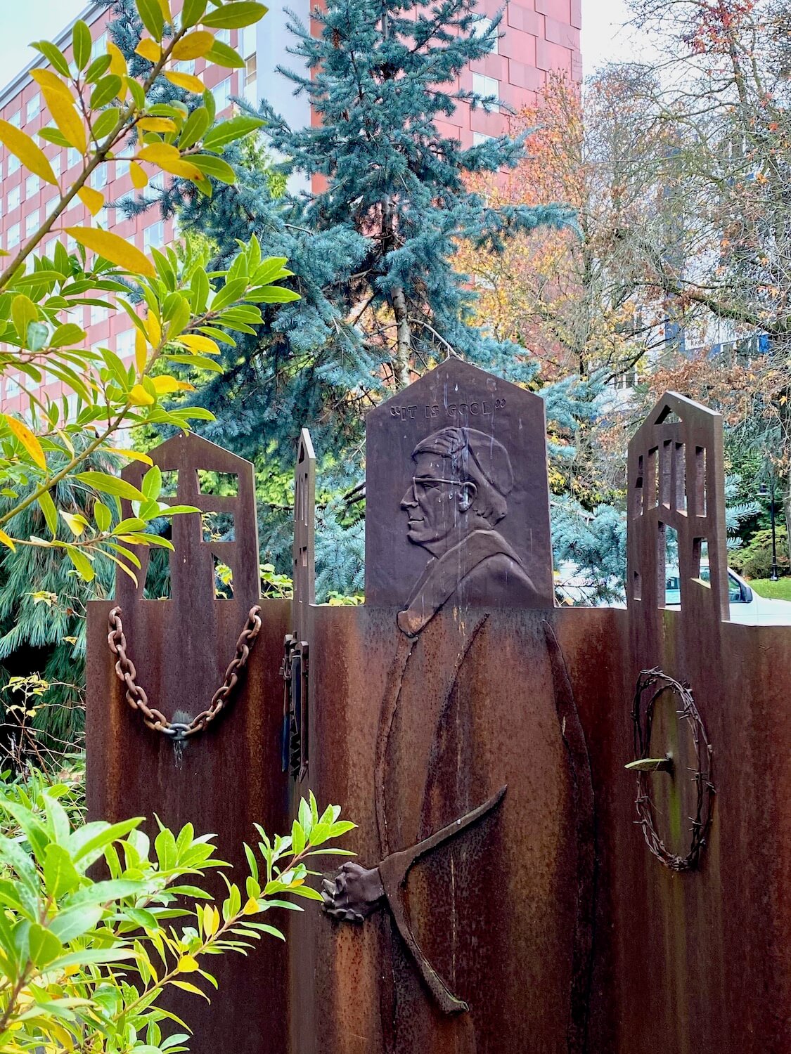 A rusted bronze art installment on the campus of Seattle University depicts a priest amongst other symbolic chunks of chain and barbed wire, making a political statement.  In the background are trees with multi-colored leaves.
