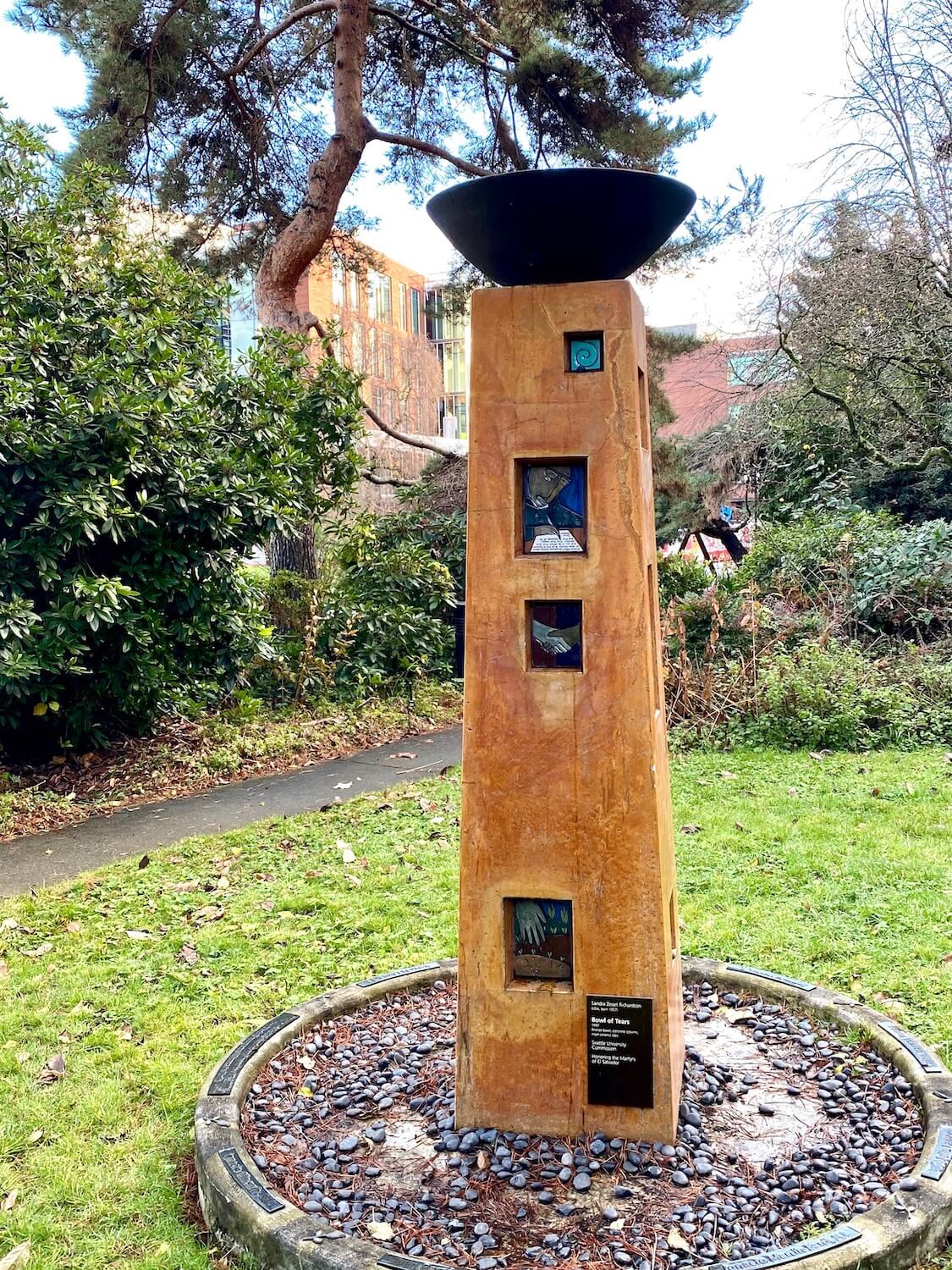 This piece of art is a column the color of orange terra cotta with squares and rectangles carved into it with tiles depicting the priests that were martyred in El Salvador.  There is a black pottery bowl on top of this column while a circle made up of stones created the base.  