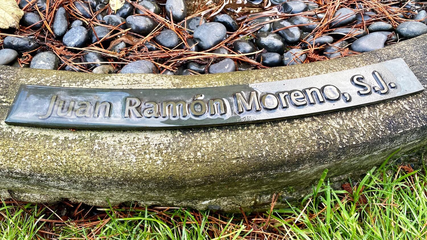 A close up of the base of the Bowl of Tears sculpture on the campus of Seattle University memorializes Juan Ramon Moreno, S.J., with a placard that is covered with water from a recent rain.  There are wet black rocks on one side of this concrete curb and grass on the other. 