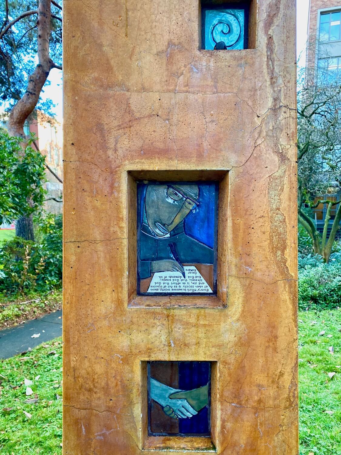 A close up of the orange terra cotta column that forms a memorial to priests martyred in El Salvador.  There is a rectangular tile embedded in the column with a priest scribing a letter.  