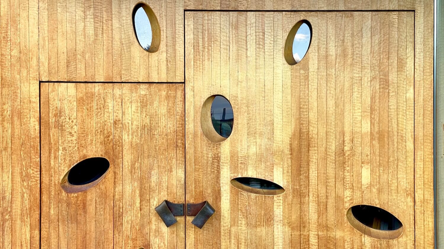 The main door of the Chapel of St. Ignatius is composed of slats of blonde wood pieced together with oblong circles cut into the wood that create windows into the church which is on the campus of Seattle University.  Two bronze door handles offer a way to open the large doors to enter the church. 