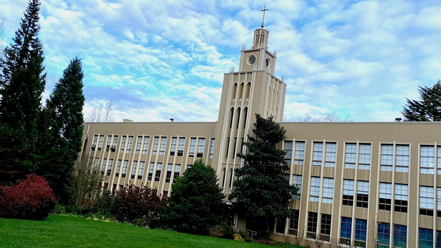 An Art Deco building on the campus of Seattle University commands a presence in front of a grassy field framed in by fir trees and a bright red shrub.  The sky is blue with scattered clouds and the cross at the top of the building seems to reach to the heavens. 