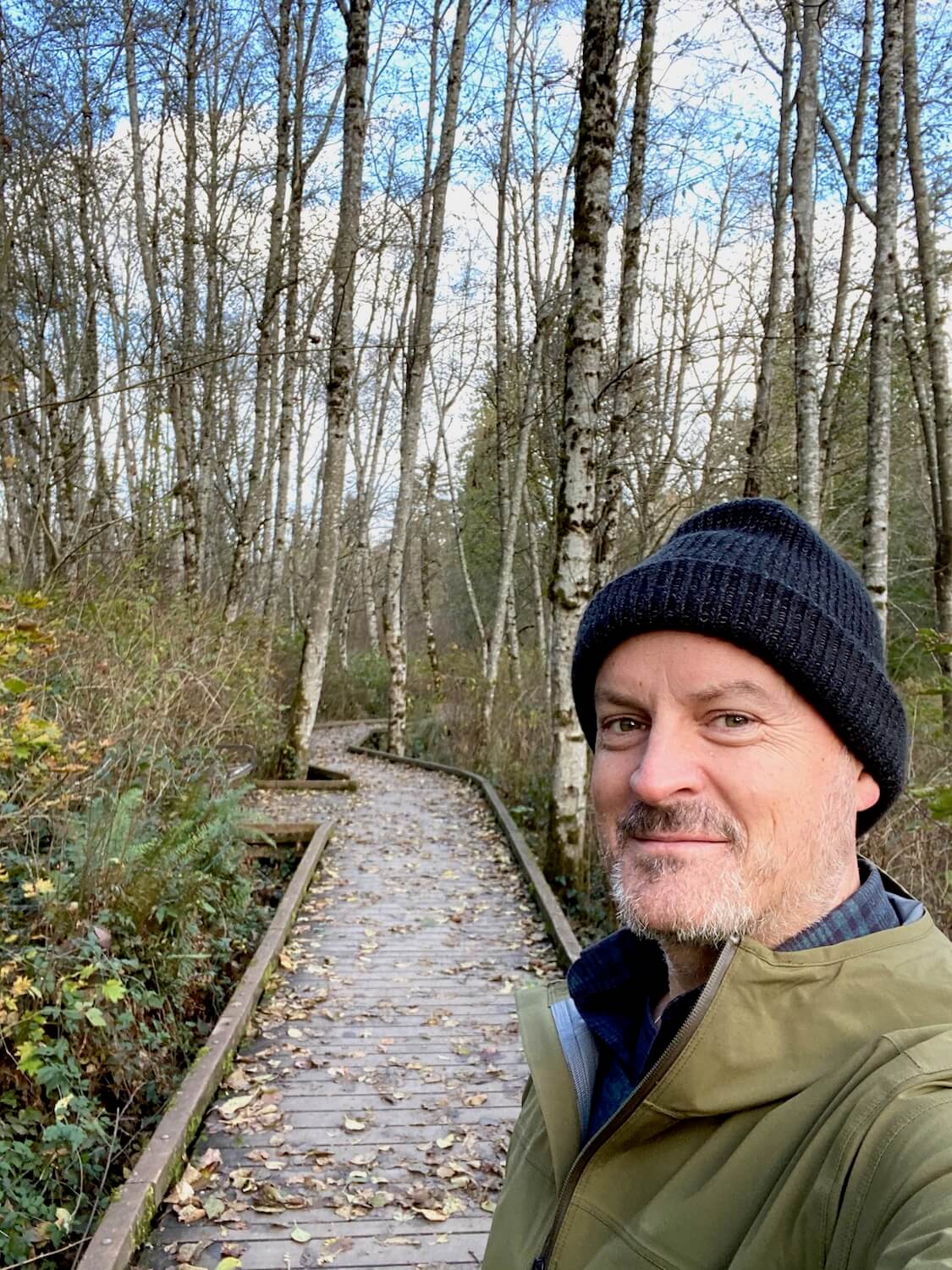 Matthew Kessi stands on a boardwalk smiling at the camera. He is wearing a black woven hat and a green rain jacket. There are birch trees in the background without leaves.