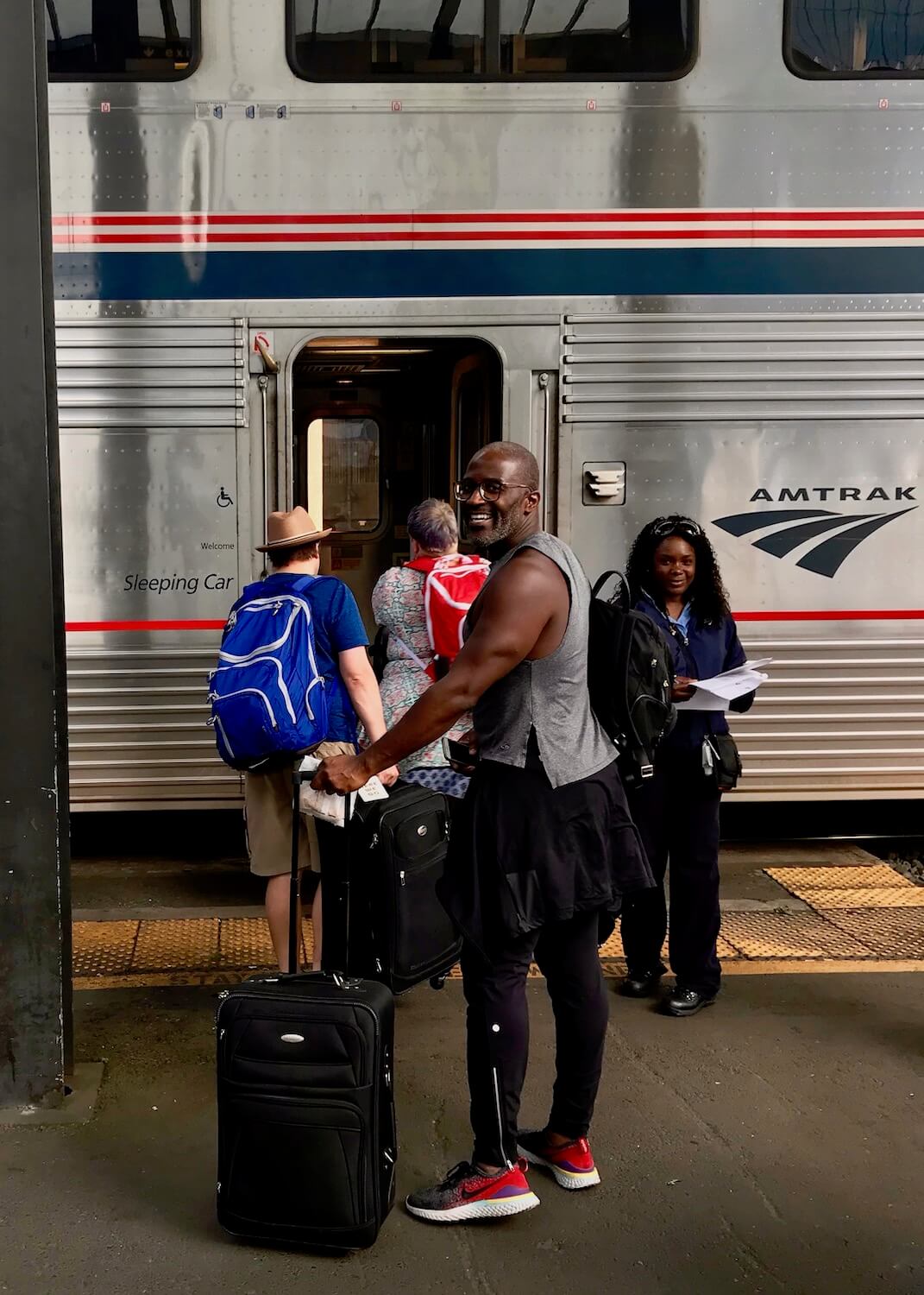 An African American man turns around and smiles at the camera while a line of passengers prepares to board a double decker Amtrak train. 