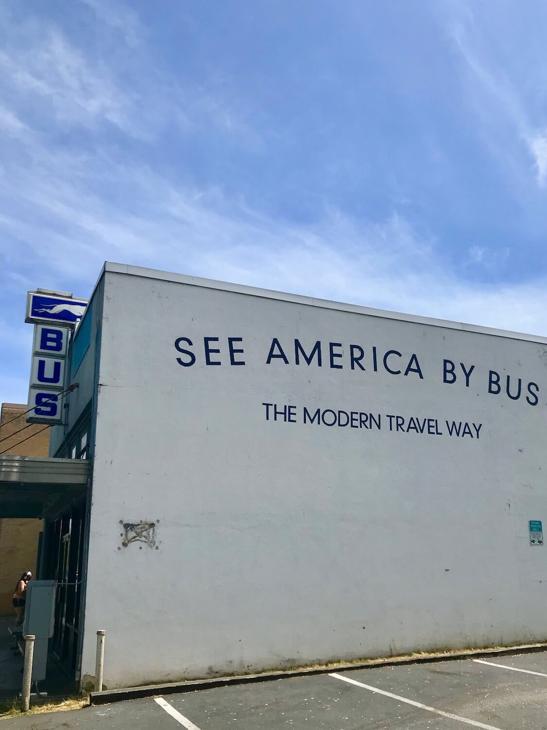 The Olympia bus station on the way to Portland from Seattle.  The blue Bus letters are topped off by the greyhound logo and the side of the building has blue letters written on the wall that say, "See America by bus."  The sky is blue with some puffy white clouds. 