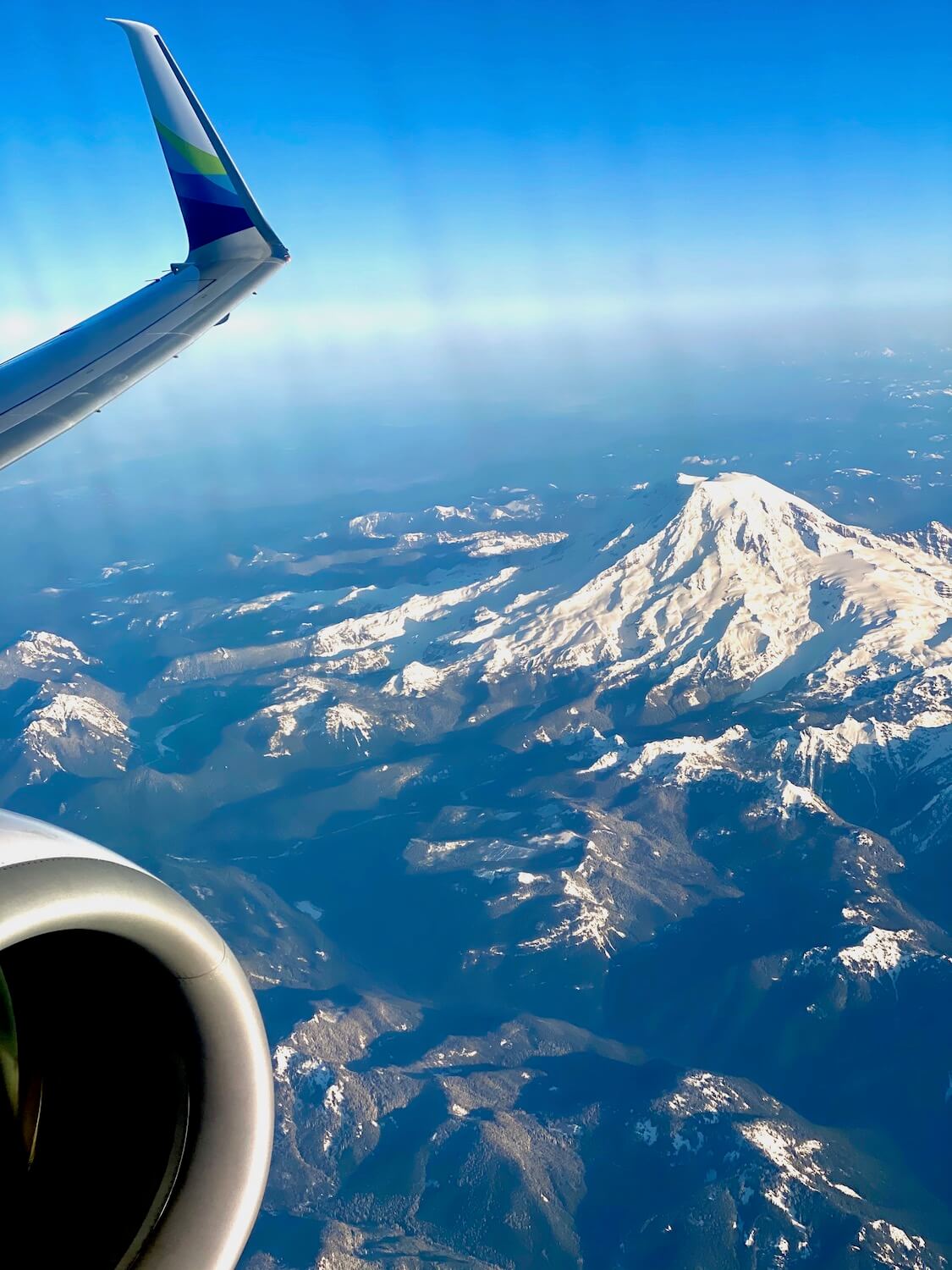 An Alaska Airlines flight to Portland from Seattle highlights majestic Mt. Rainier, covered with a heavy layer of white snow.  The winglet of the jet airplane shows the green and blues of the airline log while the sky and horizon are blue. 