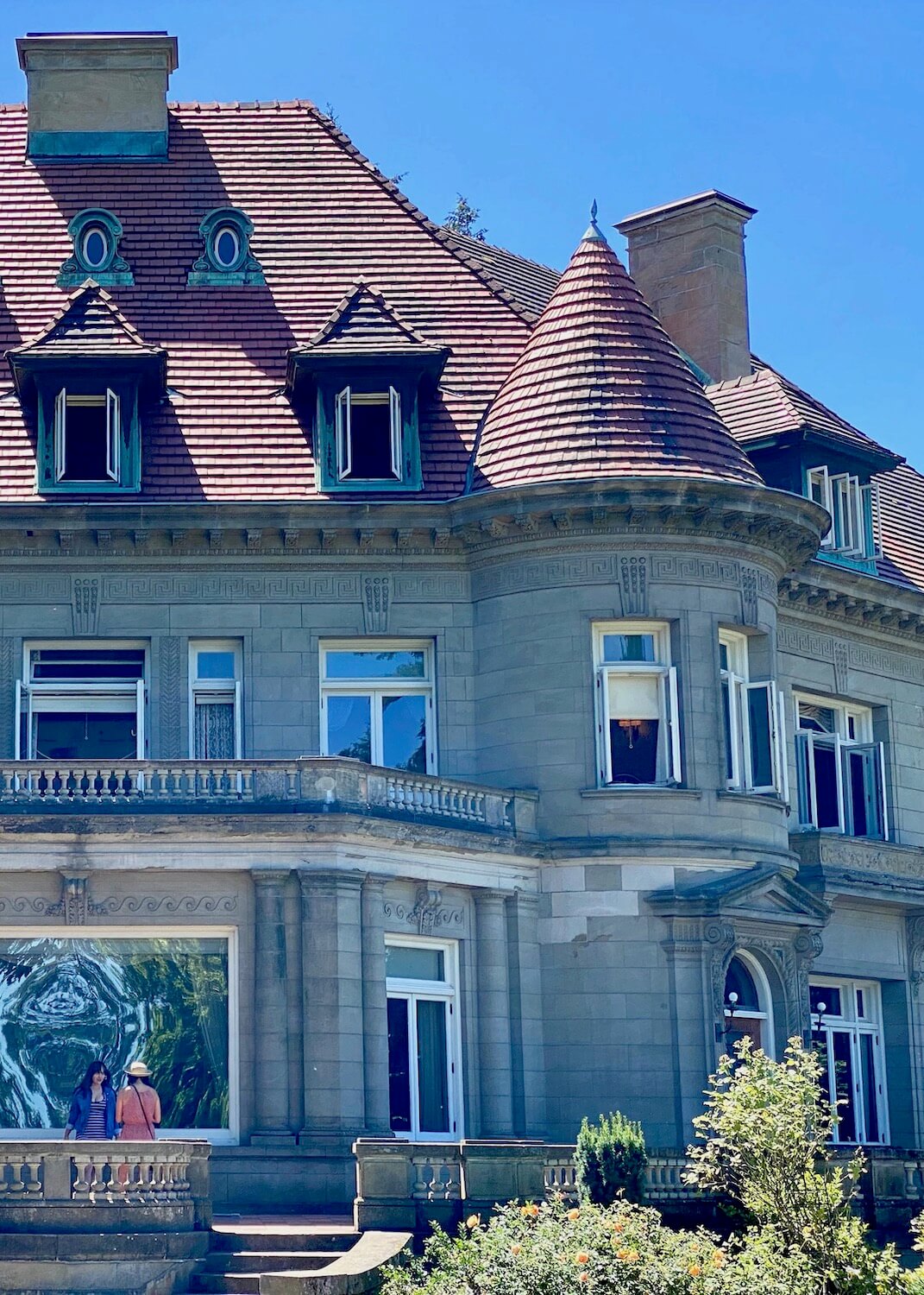 Pittock Mansion in Portland, has a prominent location overlooking the skyline of the city.  The red tiled roofs contrast with the blue sky and the heavy stone facade frames in the variety of windows, as two woman stand on the main veranda. 