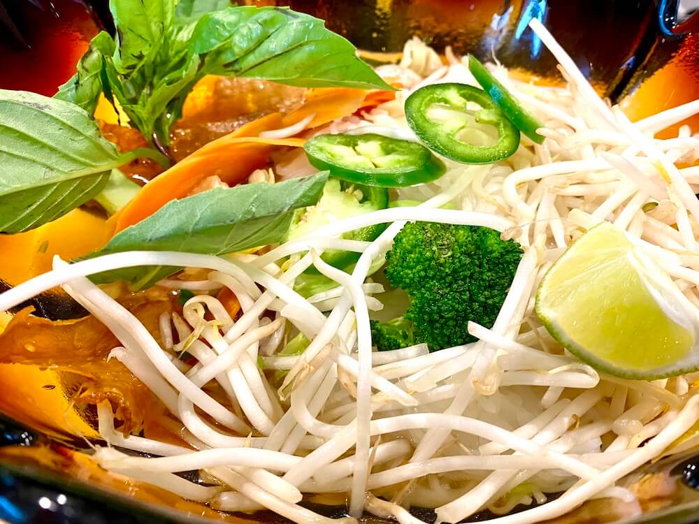 A close up shot of a bowl with tasty Pho soup that includes sliced jalapeños, basil, bean sprouts and lime.  The soup is in a orangish bowl.