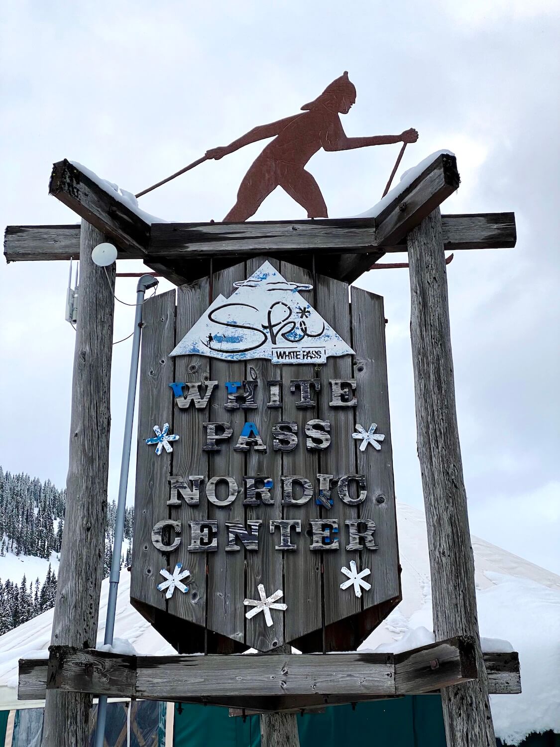 The rustic faded wood sign for White Pass Nordic Center, with gray weathered boards and the outline of a cross country skier on top.  In the back is the yurt.  