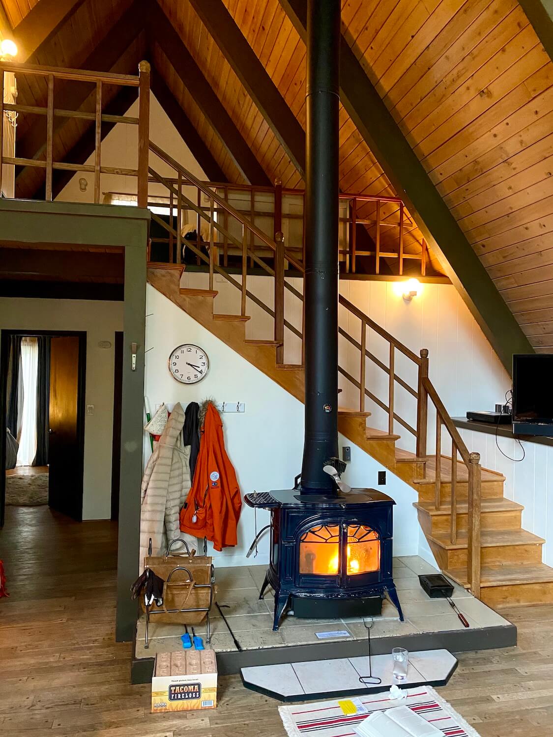 A fire roars inside a metal stove inside an a-frame cabin as wooden stairs lead up to a loft area in the space.  A clock on the wall says 3:20 and there are coats hung up on a coat rack. 