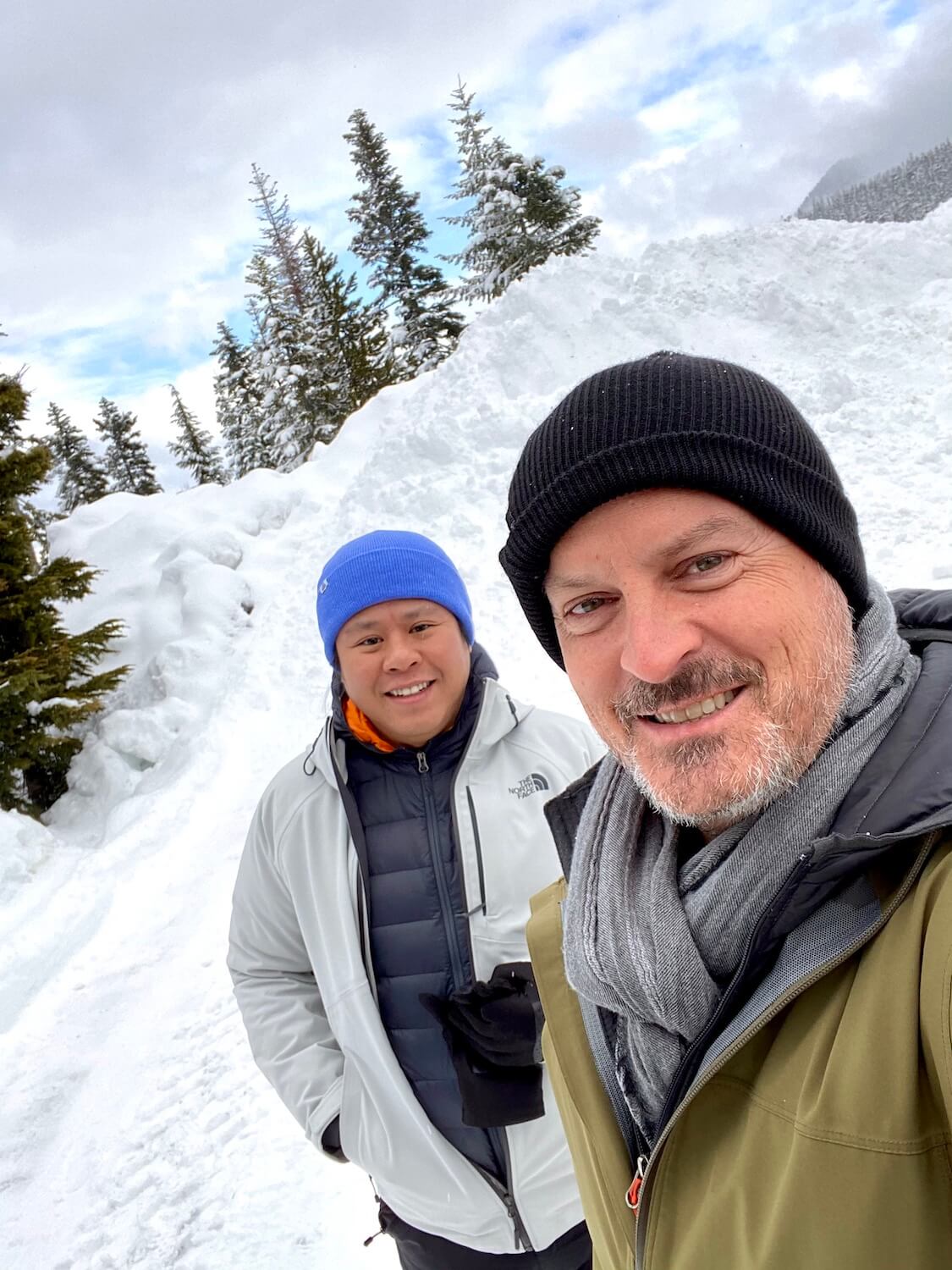 Matthew Kessi takes a selfie with his friend Mike, who is smiling and wearing a blue cap.  They are standing in front of a snow pile near the Nordic Center at White Pass and preparing to enjoy a day with snowshoes.  The blue sky is peeking through snow covered fir trees in the background.  