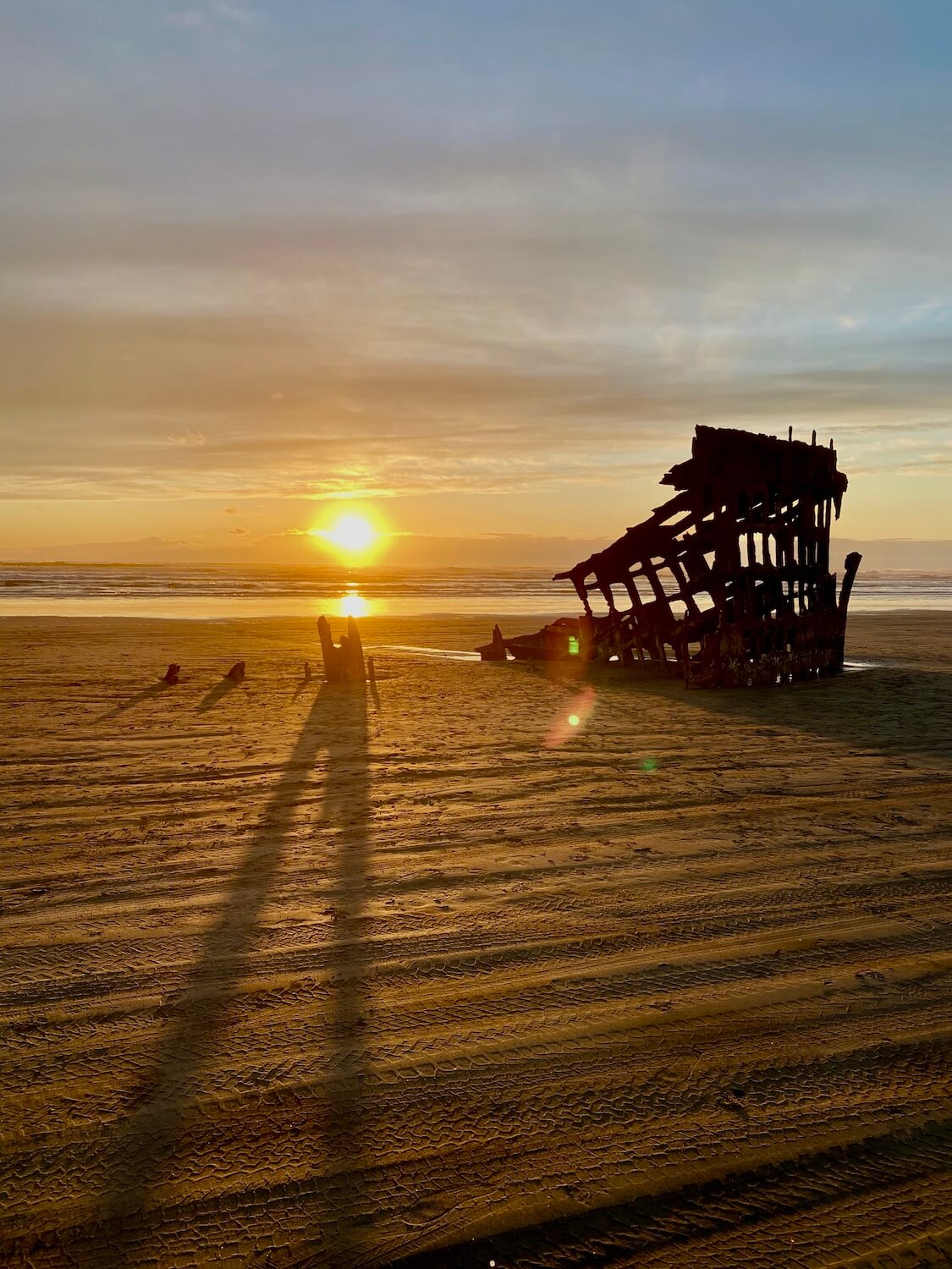 The Peter Irdale shipwreck sits wedged into the sandy beach at Fort Stevens State Park, near Astoria as the sun sets beyond the horizon of the Pacific Ocean.  The glow is yellowish as the clouds and upper sky fight to keep their blue color. 