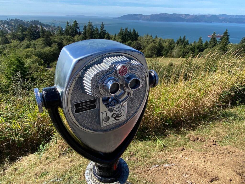 A chrome mechanical binocular will offer a closer look at the sights in the background for only 25 cents. The grass trails down to evergreen trees and eventually the waters of the Columbia River.