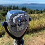A chrome mechanical binocular will offer a closer look at the sights in the background for only 25 cents. The grass trails down to evergreen trees and eventually the waters of the Columbia River.