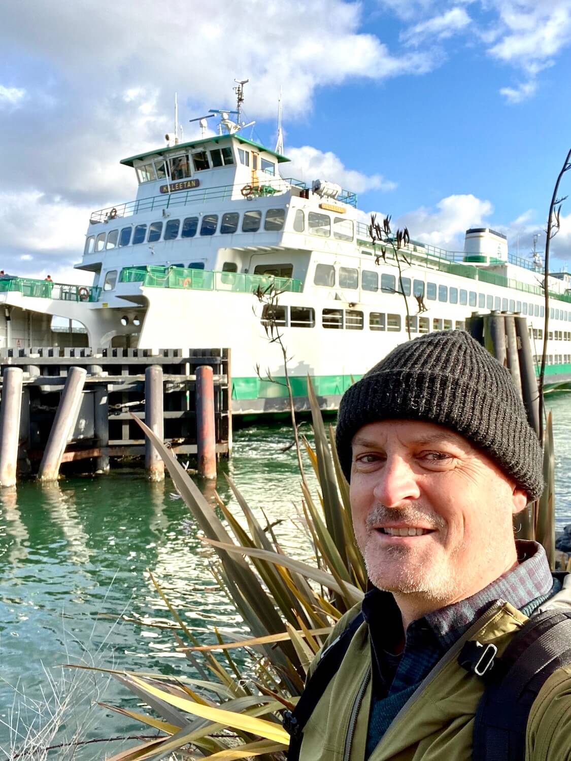 Matthew Kessi stands on the bank of the water with a Washington State Ferry in the background.  He's smiling and wearing a gray hat.  A ferry ride is a great way to spend time while on a long layover at Seatac Airport. 