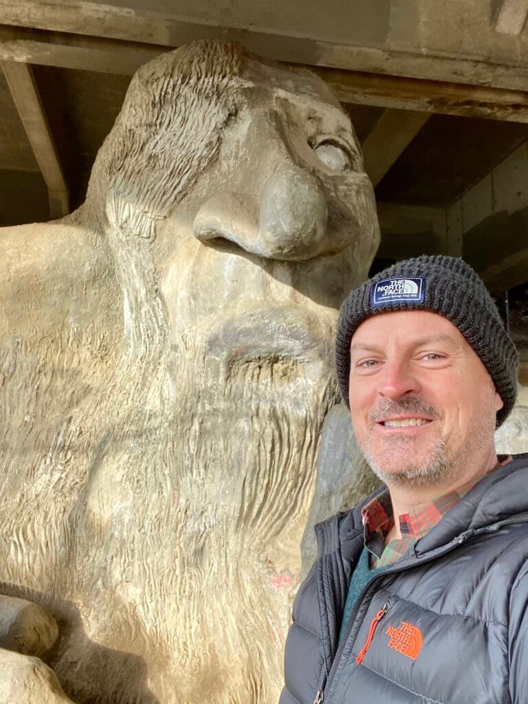 Matthew Kessi stands in front of the famous Freemont Troll, under the Aurora Bridge.  He's smiling and wearing a cap with a puffy winter coat.