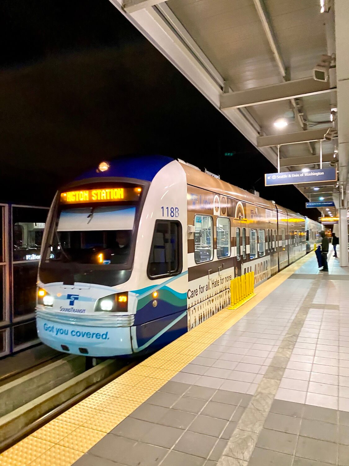 A Link Light Rail train waits at the station in a nighttime scene at Seatac Airport, preparing to head into the city center.  The train is white with a mixture of blue and green paint as well as ads on the side of the car. 