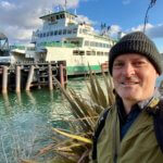 Matthew Kessi stands at the ferry dock with a green and white vessel behind him.
