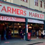 The entrance to Pike Place Market in Seattle has large neon red sign welcoming guests with brick street and cloudy sky.