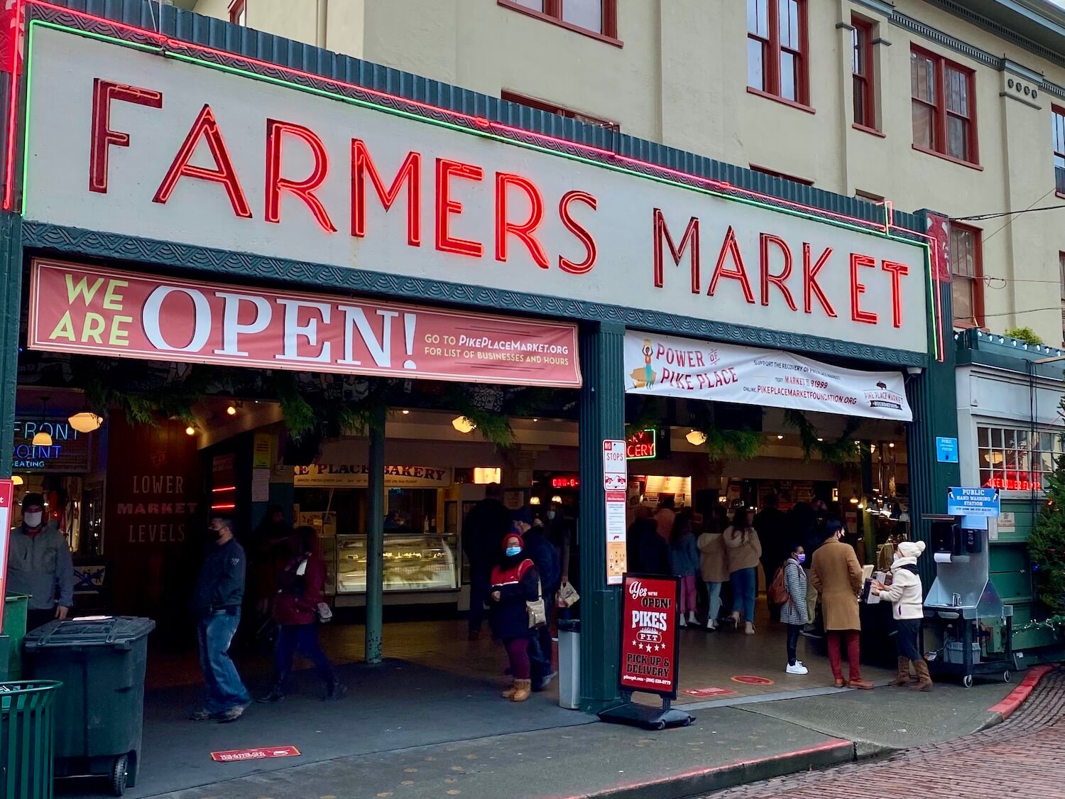 The entrance to Pike Place Market in Seattle has large neon red sign welcoming guests with brick street and cloudy sky.