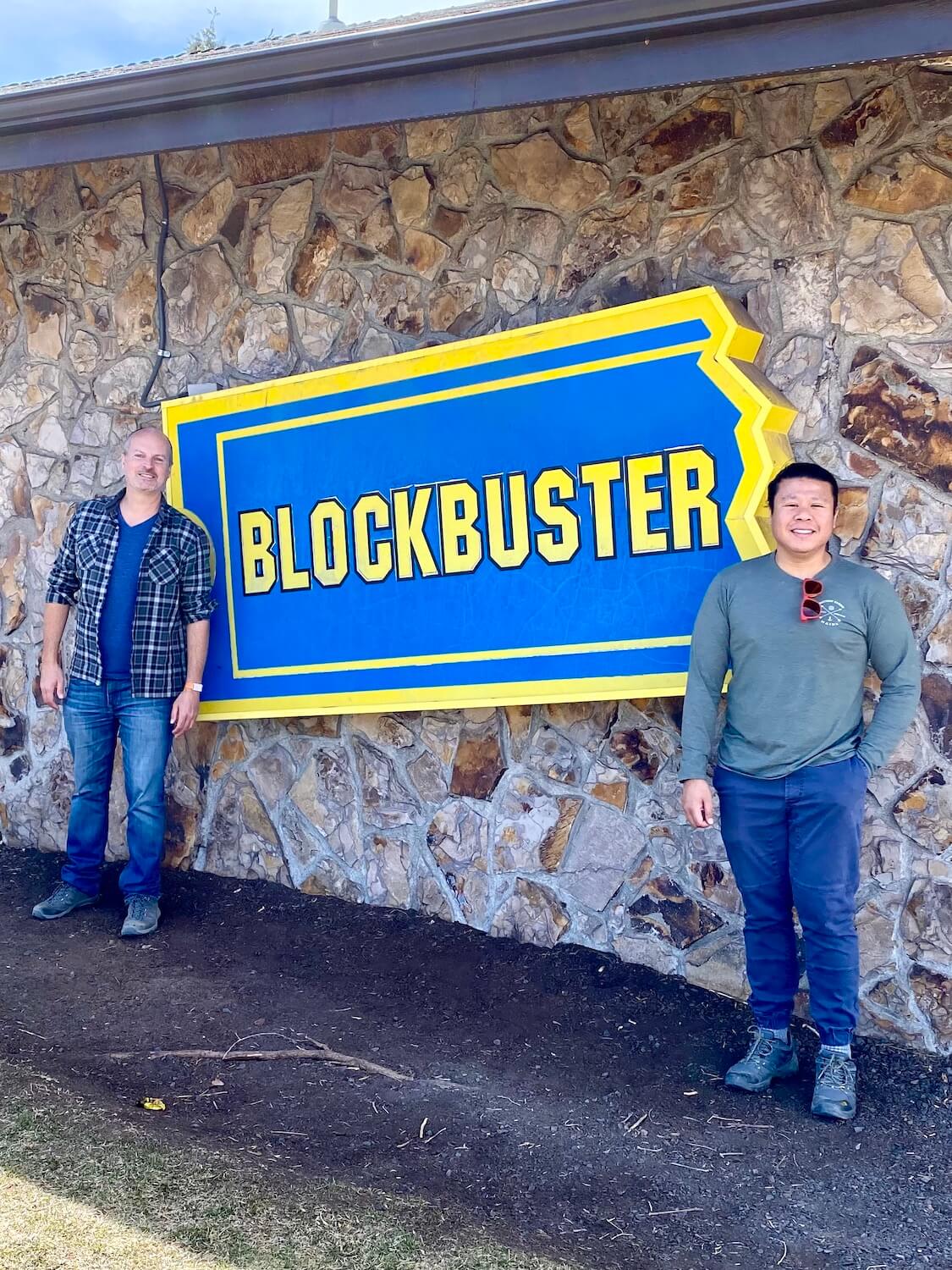 Matthew Kessi and friend stand in front of the last remaining Blockbuster Video franchise, which is in Bend, Oregon.  The large Blockbuster sign shows bright yellow lettering and blue fill in of the giant ticket stub on a brick patchwork wall. 