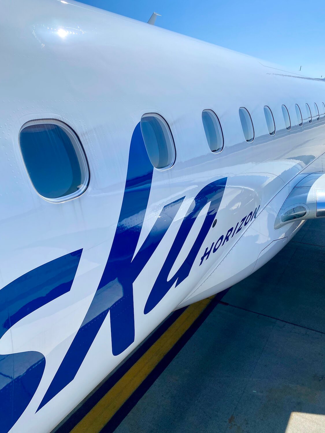 This close up shot of a Horizon Air E175 highlights the bright blue lettering on white paint and the yellow aircraft J line below is painted on the pavement.  The sky above is blue. 