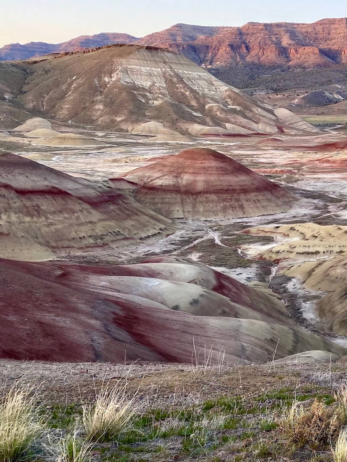 An iconic shot of the Painted Hills Oregon near sunset shows brilliant mounds of red volcanic material atop other layers of yellow and black.  The floor of this range appears to be a chalky white, while the sun still shines on a high canyon ridge in the distance. 