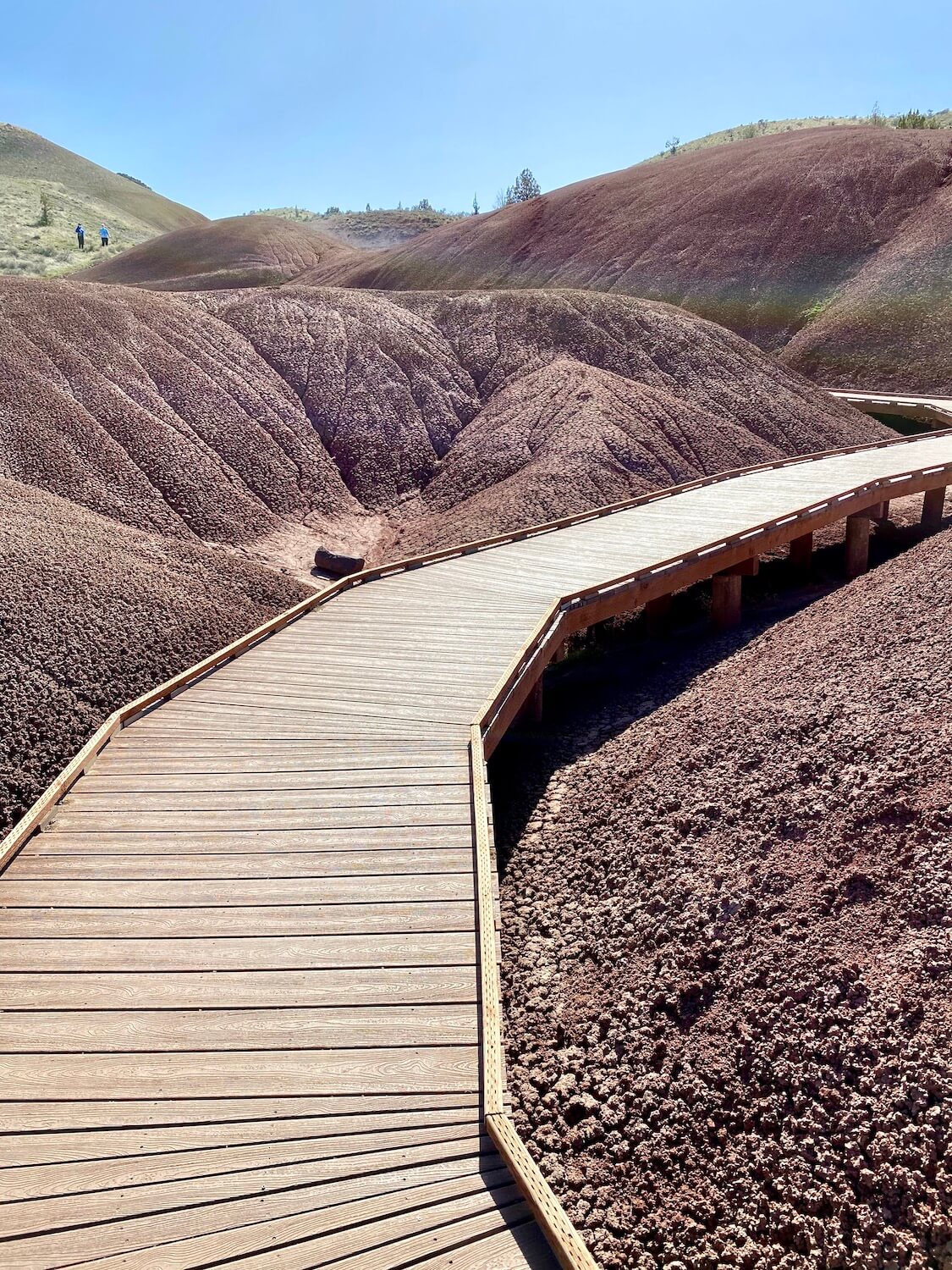 The boardwalk at the Painted Hills National Monument is a popular place for visitors because the access is so easy.  The boardwalk winds through coral like mounds of red soil while two hikers wearing blue walk toward an even larger yellow-green hill.  The sky above is bright blue and the reflection of the sun on the scene makes a light rainbow.  